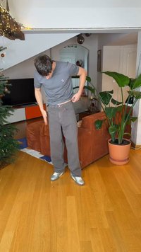 Uomo con t-shirt grigia e pantaloni che sistema la cintura nel soggiorno con divano marrone, grande pianta e albero di Natale con luci.