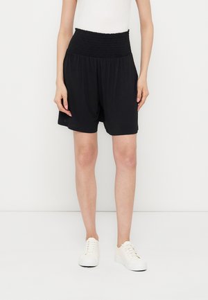 Kobieta ubrana w czarne szorty z wysokim stanem i marszczonym paskiem, biały top bez rękawów i białe trampki, stojąca na białym tle.