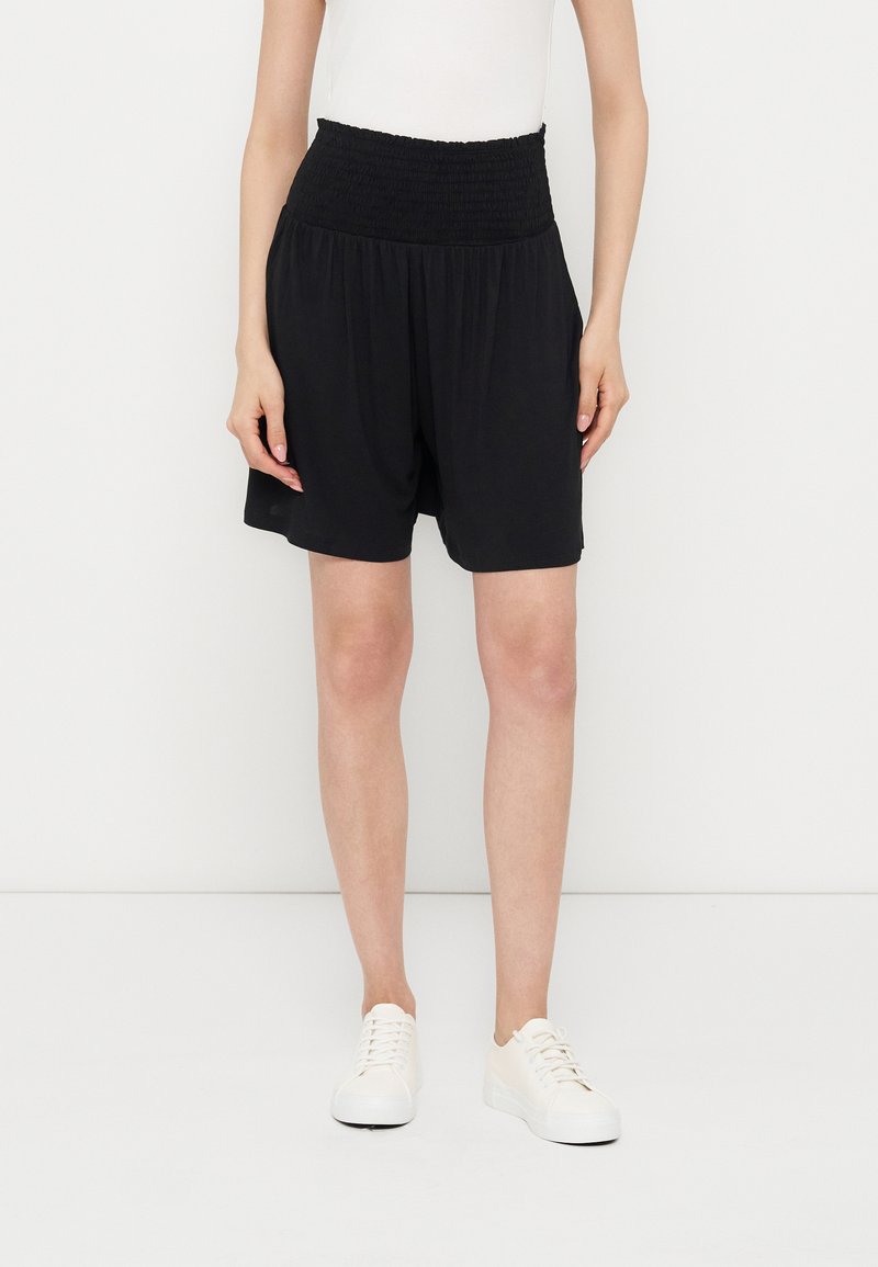 Γυναίκα με μαύρο σορτς ψηλόμεσο με ελαστική μέση, άσπρο αμάνικο τοπ και λευκά αθλητικά παπούτσια, που στέκεται σε λευκό φόντο.