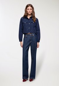 Giacca cropped trapuntata blu navy con due tasche sul petto, abbinata a jeans in denim scuro a gamba larga con vita alta e calzature rosse lucide.