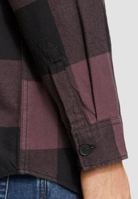Chemise à manches longues à carreaux noir et bordeaux, avec un poignet à boutons et un tissu texturé présentant des détails de couture visibles.