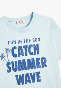 Lyseblå bomulds T-shirt med korte ærmer. Har stor, fed, blå tekst: "SJOV I SOLEN TAG SOMMERBØLGEN," og grafik af palmetræer.
