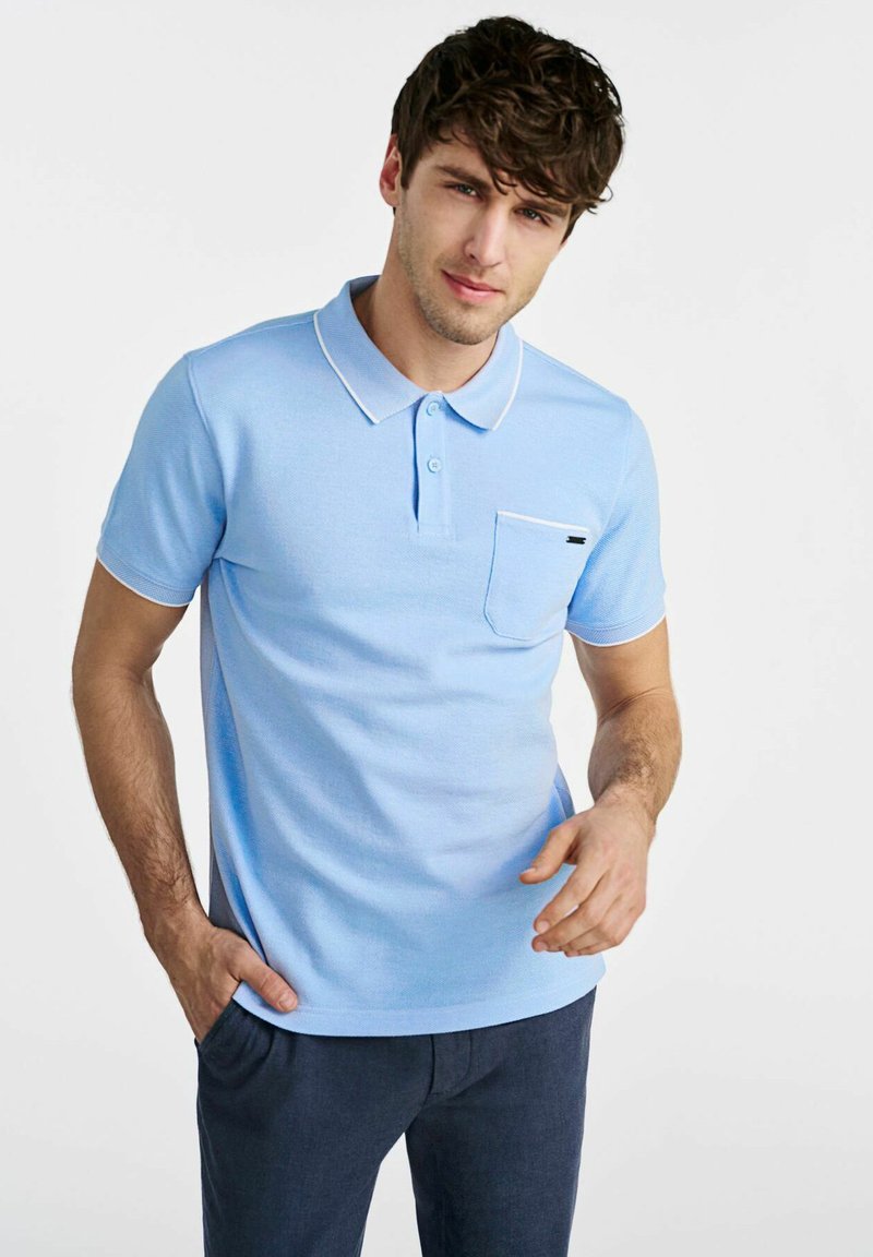 Jasnoniebieska koszula polo z teksturowanym wzorem, z trzema guzikami, białym kołnierzykiem i wykończeniem kieszeni, z małym czarnym akcentem na kieszeni.
