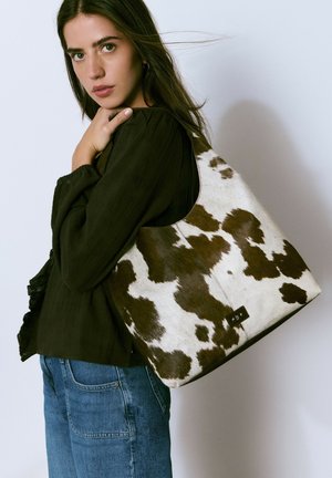 Bolso tipo hobo hecho de cuero de vaca con patrones marrones y blancos, que presenta una textura suave y una etiqueta rectangular negra.