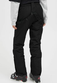 Black ski pants feature a relaxed fit, elastic waistband, and a printed strap along the back. Paired with black ski boots.
