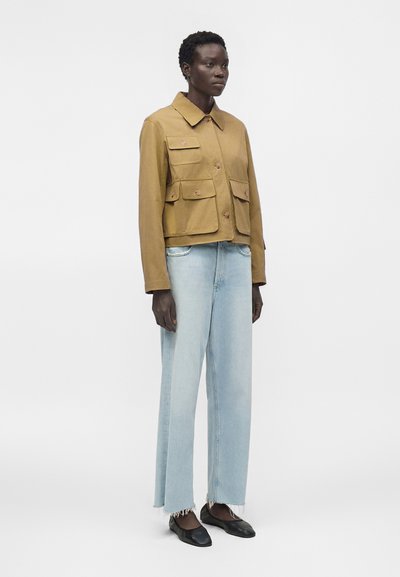 Άτομο ντυμένο με μπεζ κουμπωτό σακάκι με τέσσερις τσέπες με καπάκια, ανοιχτό μπλε φαρδύ τζιν με ξεφτισμένα τελειώματα και μαύρα επίπεδα παπούτσια, που στέκεται πλάγια.