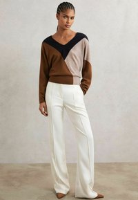 Pullover a V in color block nei toni marrone, beige e blu navy, abbinato a pantaloni bianchi a vita alta; texture liscia e vestibilità sartoriale.