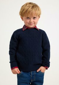 Granatowy sweter w warkoczowy wzór z prążkowanymi mankietami i dołem, nałożony na czerwono-szaro-kraciastą koszulę z kołnierzykiem. Chłopiec stoi ze dłońmi w kieszeniach, w niebieskich dżinsach.