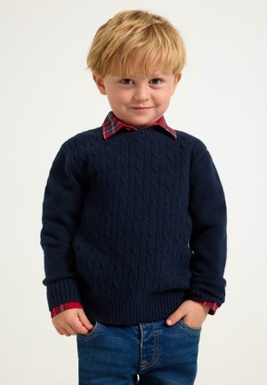 Maglione navy a lavorazione a trecce con polsini e orlo a costine, indossato sopra una camicia a righe rosse con colletto. Ragazzo in piedi con le mani in tasca, indossa jeans blu.