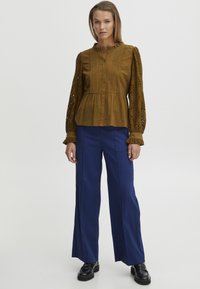 Blusa peplum in senape con chiusura a bottone, colletto arricciato e maniche in pizzo con motivo elaborato. Abbinata a pantaloni a gamba larga blu navy.