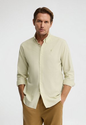 Man met lichtgroen gestreept overhemd met opgerolde mouwen en een bruine broek, staand met handen in de zakken tegen een witte achtergrond.