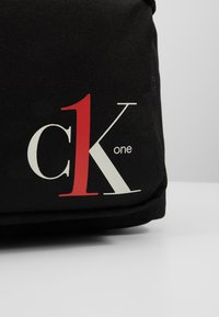 Czarna torba z tkaniny z wyraźnym czerwono-białym logo "ck one" z jednej strony, o fakturowanej powierzchni i zaokrąglonych krawędziach.