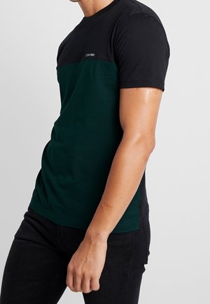 Homme portant un t-shirt à manches courtes Calvin Klein en noir et vert foncé à blocs de couleur, et un pantalon noir, debout devant un fond uni.