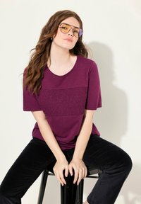 Camisa morada con panel texturizado, de mangas cortas; combinada con pantalones de pana negros. El modelo lleva gafas de sol con tintado naranja, sentado en un taburete negro.
