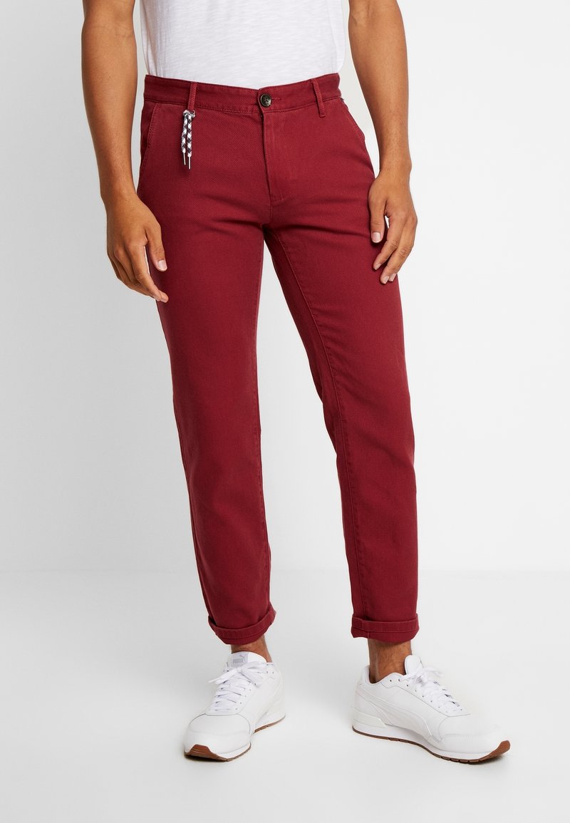 Man in slim-fit donkerrode broek met opgerolde pijpen, witte sneakers en een wit T-shirt, staand tegen een effen lichte achtergrond.