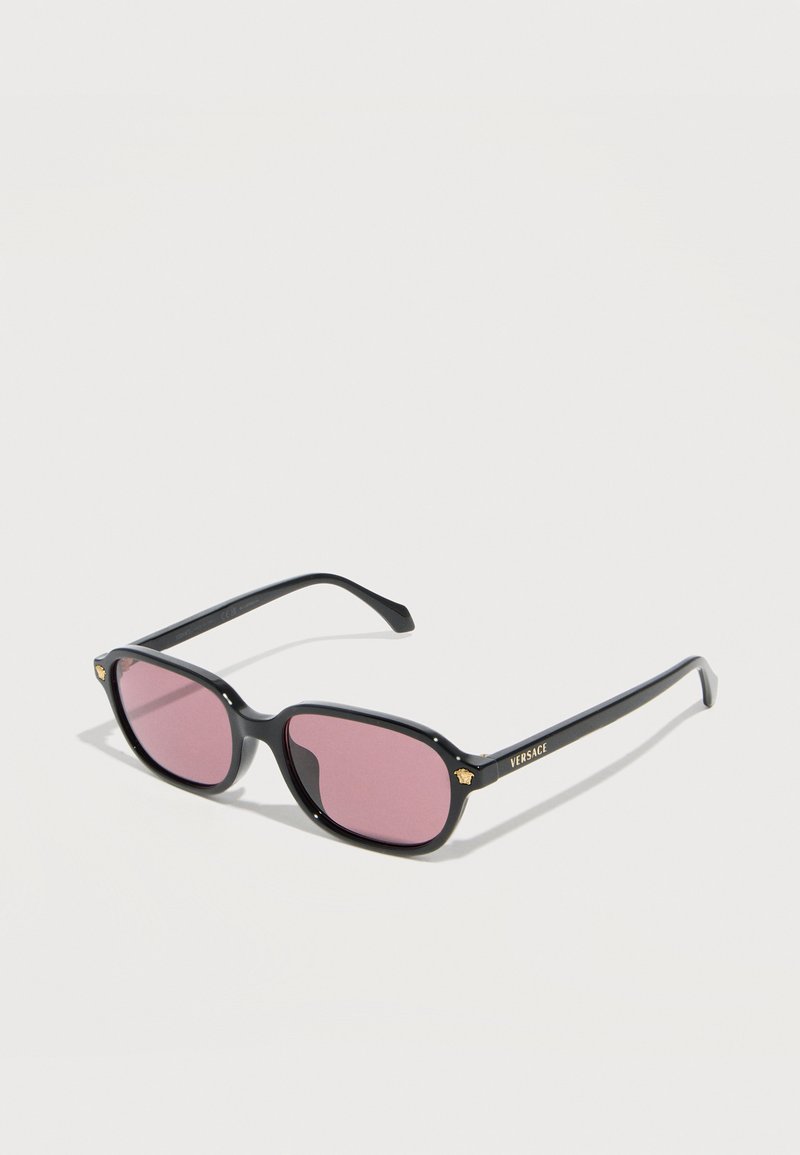 Μαύρα ορθογώνια γυαλιά ηλίου με ροζ φακούς, χρυσά λογότυπα Medusa στους βραχίονες και τη σήμανση "Versace" στο δεξί μπράτσο, εκτεθειμένα σε λευκή επιφάνεια.