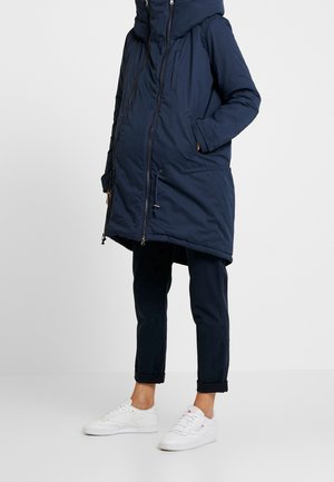 Persoon die een marineblauwe gewatteerde jas met zakken draagt, navy opgerolde broek en witte sneakers, staand tegen een effen achtergrond.