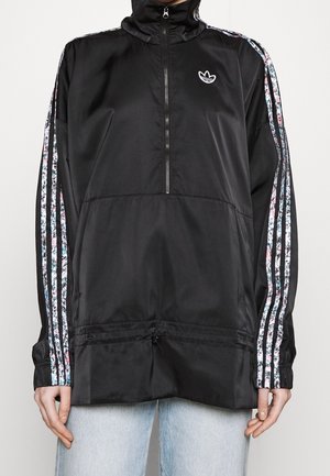 Veste adidas noire à demi-fermeture éclair avec trois bandes à motif floral sur les manches et logo sur la poitrine, portée sur un jean bleu clair.