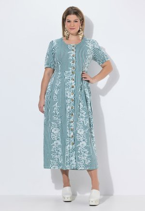 Vrouw die een midi-jurk draagt met groene en witte strepen en bloemmotieven, knopen aan de voorkant, korte mouwen, gouden opvallende oorbellen en witte plateau schoenen.