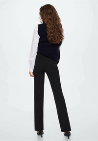 Black fitted pants paired with a dark blue knit vest over a white long-sleeve shirt, viewed from the back. Long, wavy hair is visible.