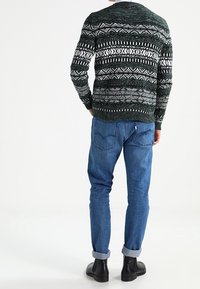 Suéter verde con patrón de diseños geométricos blancos, textura tejida. Combinado con jeans azules y botas negras hasta el tobillo, mostrando un look casual.