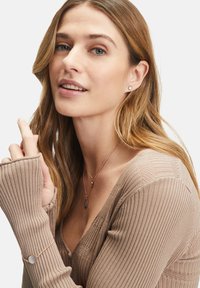 Pull à côtes beige avec manches longues. Présente des colliers en couche en ton doré avec un pendentif ovale et des boucles d'oreilles à clou. Textures lisses évidentes.