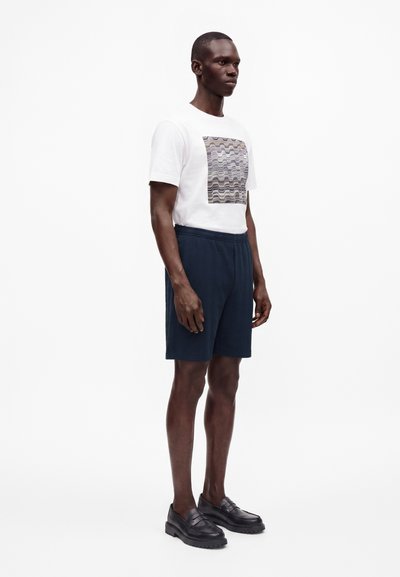 Homme debout portant un t-shirt blanc à motif graphique, un short texturé bleu marine et des mocassins noirs sur un fond blanc uni.