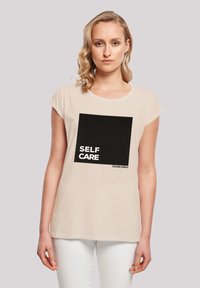 Beige Baumwoll-T-Shirt mit kurzen Ärmeln, das ein schwarzes quadratisches Grafikdesign zeigt, auf dem "SELF CARE" in weißem Text steht. Schlichte, lässige Passform.