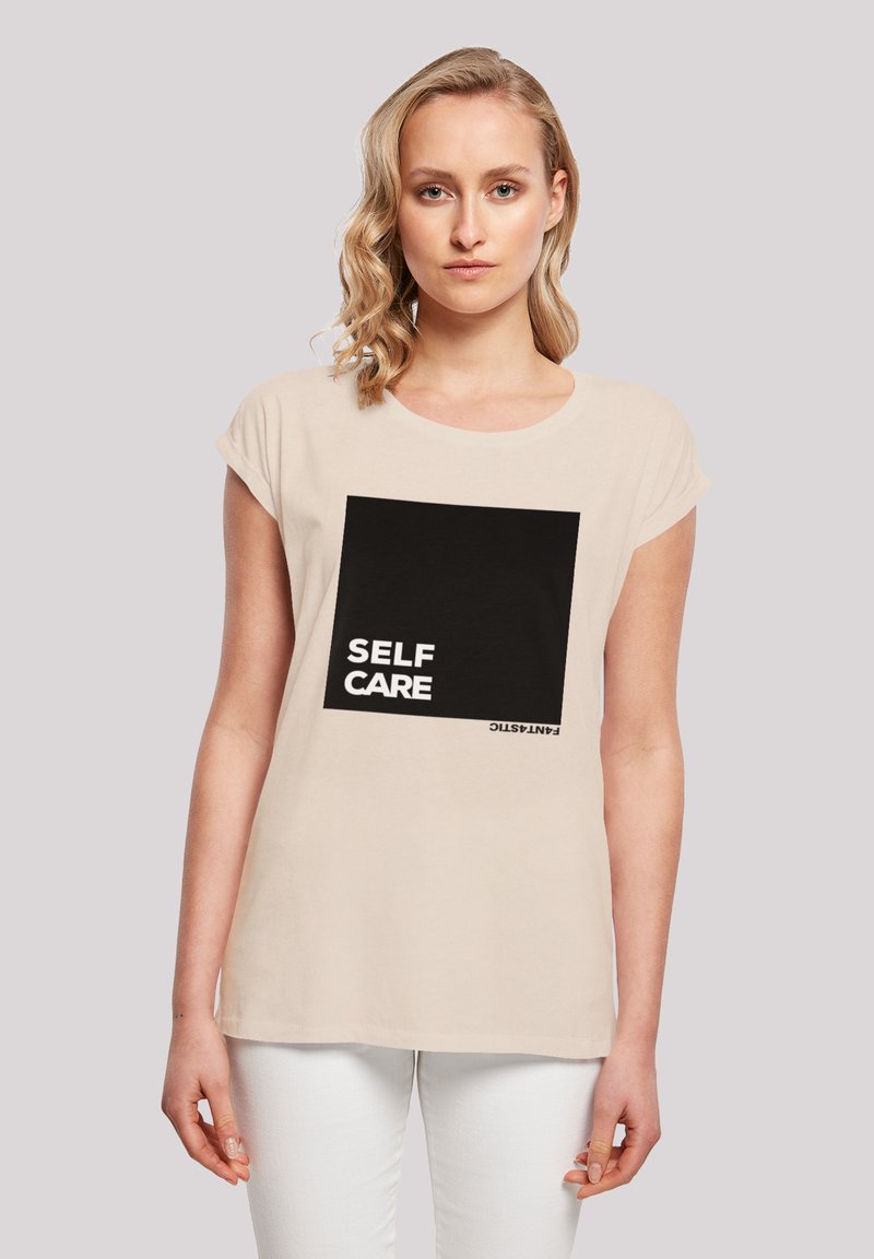 Beige Baumwoll-T-Shirt mit kurzen Ärmeln, das ein schwarzes quadratisches Grafikdesign zeigt, auf dem "SELF CARE" in weißem Text steht. Schlichte, lässige Passform.