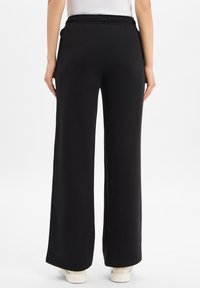 Kobieta w czarnych szerokich spodniach i białych trampkach, stojąca na tle jednolitej białej ściany, widziana z tyłu.