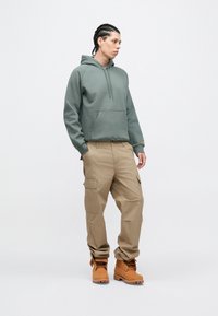 Zelena majica s kapuljačom s prednjim džepom, bež cargo hlače s više džepova i smeđe gležnjače sa vezicama; ležerni, opušteni stil.