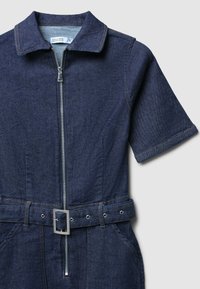 Salopette en denim avec col, manches courtes, fermeture éclair à l'avant, taille ceinturée avec une boucle en argent et d'une couleur bleu foncé.