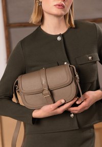 Beige tekstureret læder skuldertaske med klaplukning, detaljer i guld og aftagelig rem, holdt mod et mørkegrønt outfit.