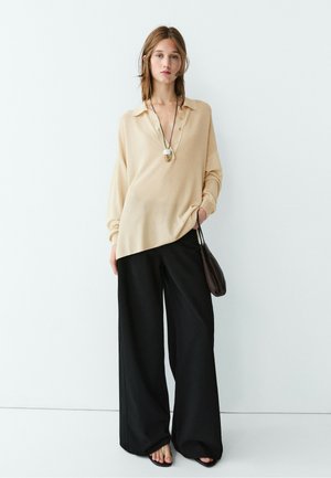 Frau trägt beige langärmelige Bluse, schwarze weite Hose, schwarze Sandalen und eine runde Anhänger-Halskette, hält eine kleine schwarze Handtasche.