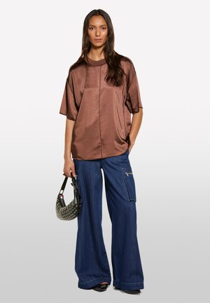 Top corto in satin marrone a maniche corte con scollatura rotonda, abbinato a pantaloni in denim scuro a gamba larga. Il modello tiene in mano una borsa a tracolla nera con motivo.