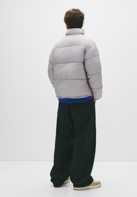 Lys grå dunjakke med høj krave, quiltet design og en afslappet pasform, kombineret med mørkegrønne brede bukser og beige sneakers.