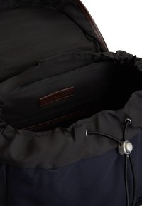 Εσωτερικό σακιδίου πλάτης από ύφασμα ναυτικού μπλε με κλείσιμο με κορδόνι, που διαθέτει τσέπη με φερμουάρ και δερμάτινη ετικέτα. Ομαλή υφή, μινιμαλιστικός σχεδιασμός.