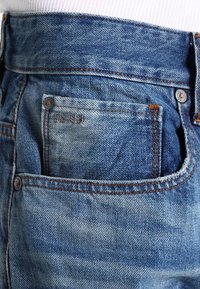 Modré džínové kalhoty s klasickým střihem, viditelným šitím a malou přední kapsou s vybledlou oblastí, doplněné bronzovým knoflíkem.