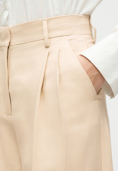Pantalon beige plissé taille haute avec passants pour ceinture, poches latérales, porté avec un haut blanc à manches longues, main dans la poche.