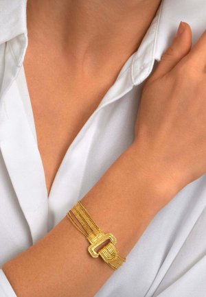 Poignet de femme portant un bracelet en or multi-brins avec un fermoir rectangulaire texturé sur une chemise blanche à poignets.