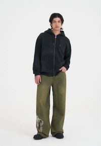 Černá mikina s kapucí na zip a přední kapsou, spárovaná s volnými olivově zelenými kalhotami, které mají grafický design a složité švy.