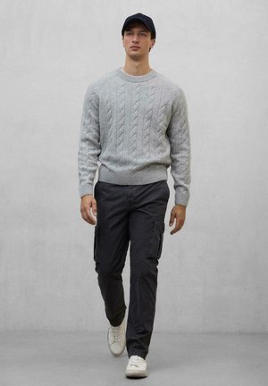 Suéter de punto trenzado en gris claro con cuello redondo, combinado con pantalones cargo oscuros y zapatillas blancas. El modelo lleva una gorra negra.