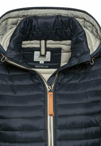 Navy blauwe pufferjack met een beige voering, gequilt textuur, zilveren rits en een bruin leren trekker. Heeft een label voor gerecyclede materialen.