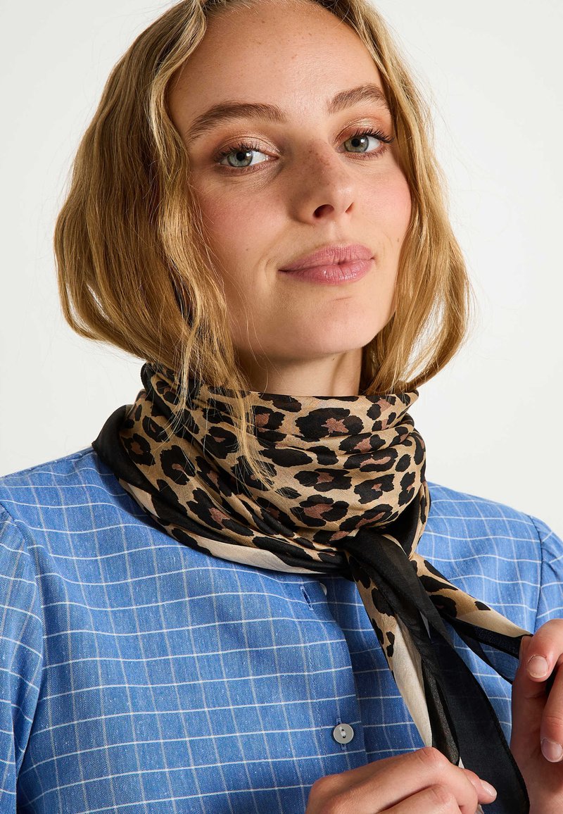 Écharpe en soie imprimée léopard avec une palette de couleurs beige et noire, présentant une texture lisse et nouée élégamment autour du cou.