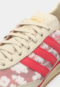 Adidas sneakers har en kremfarget overdel, rosa striper, teksturert blomsterdesign og beige lisser. Merkelig gulllogo på tungespissen.