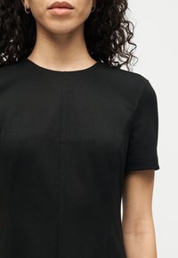 Γυναίκα με κατσαρά μαλλιά που φοράει μαύρο φόρεμα με κοντά μανίκια και μικρούς κρίκους σκουλαρίκια, εμφανίζεται από τους ώμους έως τα χείλη σε λευκό φόντο.