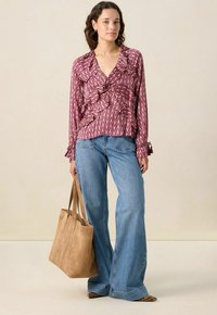 Frau mit einer lila gemusterten Bluse, weit geschnittenen blauen Jeans, die eine große hellbraune Wildleder-Tasche hält, vor einem neutralen Hintergrund stehend.