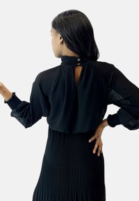 Vestido negro plisado con mangas largas y transparentes, cuello alto con detalle de botones y un diseño de espalda con ojal. La tela texturizada añade dimensión.