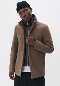 Abrigo marrón de mezcla de lana con un alto cuello, cremallera de largo completo y bolsillos laterales, superpuesto a un abrigo de plumas azul marino. Gorro de punto negro en el modelo.