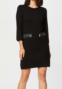 Robe noire à manches trois-quarts, avec deux poches à glissière argentées sur le devant. Texture lisse et design à coupe droite.